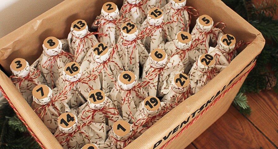 Ein Holzkasten mit der Aufschrift 'ADVENTSKALENDER' enthält 24 in Geschenkpapier verpackte Flaschen, nummeriert von 1 bis 24. Der Kasten ist mit Tannenzweigen und Weihnachtsdekoration geschmückt.