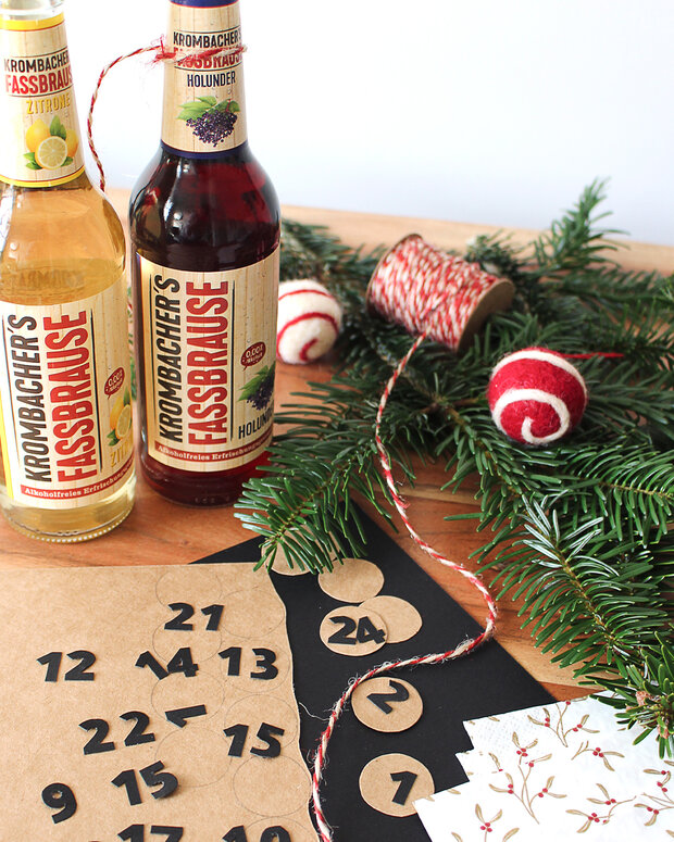 Zwei Flaschen Krombacher's Fassbrause in den Geschmacksrichtungen Zitrone und Holunder stehen auf einem Tisch. Daneben liegen dekorative Elemente wie eine Tannenzweige, eine Schnur und ein Adventskalender mit Zahlen.