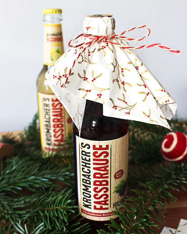 Zwei Flaschen Krombacher's Fassbrause stehen auf einem Tisch, eine ist mit einem dekorativen, gemusterten Tuch bedeckt. Die Flasche im Vordergrund zeigt ein Etikett mit dem Schriftzug 'Krombacher's Fassbrause'.