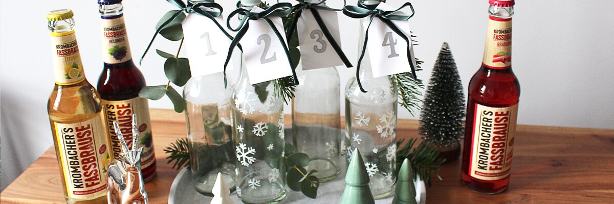 Fünf Flaschen mit grünen Kerzen und nummerierten Anhängern stehen auf einem runden Tablett. Links zwei Flaschen 'Krombacher's Kasselbrause' und rechts eine Flasche 'Krombacher's Kasselbrause' in rot.