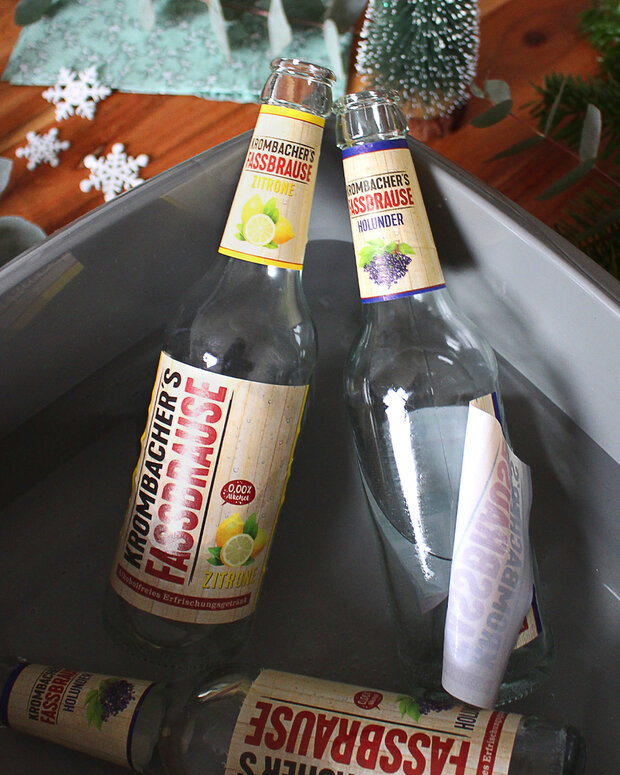 Zwei Flaschen Ironbacher's Froschbrause in einem grauen Behälter, eine mit Zitrone und die andere mit Holunder. Beide Flaschen sind teilweise mit Etiketten sichtbar.