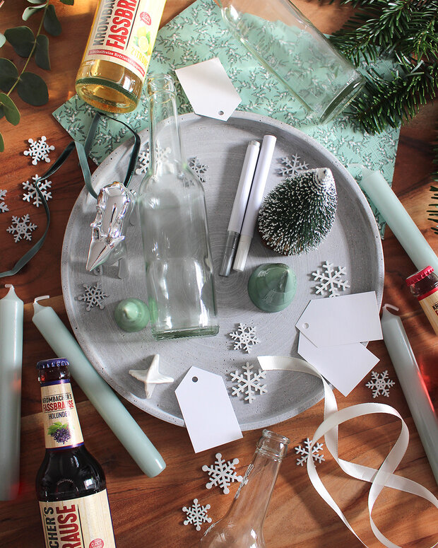 Ein grauer Teller mit leeren Glasflaschen, grünen Kerzen, Schneeflocken und Geschenkanhängern. Dekorative Elemente wie ein kleiner Tannenbaum und eine Flasche Fassbrause sind ebenfalls sichtbar.