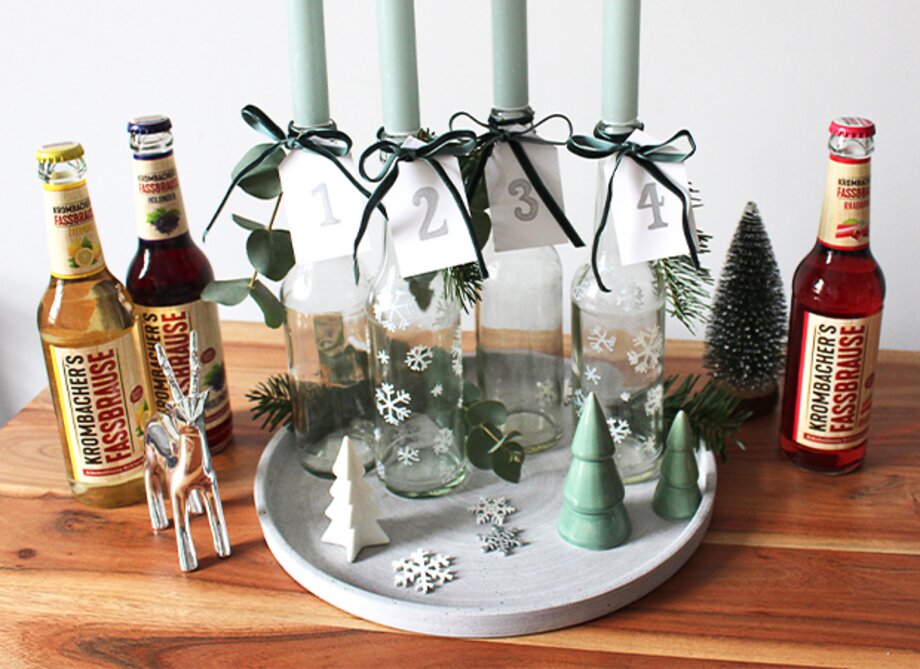 Fünf Flaschen mit grünen Kerzen und nummerierten Anhängern stehen auf einem runden Tablett. Links zwei Flaschen 'Krombacher's Kasselbrause' und rechts eine Flasche 'Krombacher's Kasselbrause' in rot.